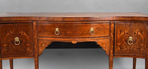 fig 2. An antique mahogany serpentine sideboard featuring floral marquetry inlays, a central drawer, two side cupboards, tapered square legs, and brass lion-mask ring handles.