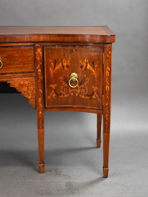 fig 3. An antique mahogany serpentine sideboard featuring floral marquetry inlays, a central drawer, two side cupboards, tapered square legs, and brass lion-mask ring handles.