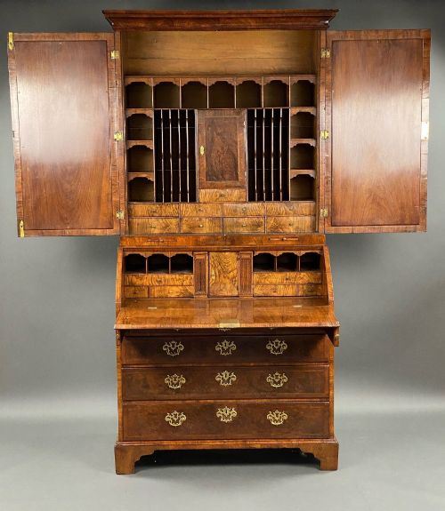 fig 1. A George II burr walnut bureau bookcase featuring an upper double-door cabinet with shaped mirrored panels over a fall-front desk and four drawers with brass handles.