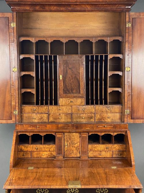 fig 2. A George II burr walnut bureau bookcase featuring an upper double-door cabinet with shaped mirrored panels over a fall-front desk and four drawers with brass handles.