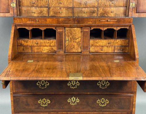 fig 4. A George II burr walnut bureau bookcase featuring an upper double-door cabinet with shaped mirrored panels over a fall-front desk and four drawers with brass handles.