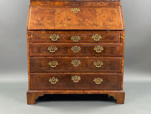fig 6. A George II burr walnut bureau bookcase featuring an upper double-door cabinet with shaped mirrored panels over a fall-front desk and four drawers with brass handles.