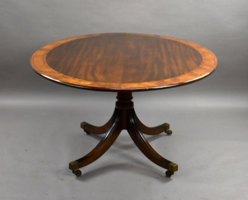 fig 1. An oval Regency style flame mahogany tilt-top dining table featuring a crossbanded satinwood border and a pedestal base with brass castors.