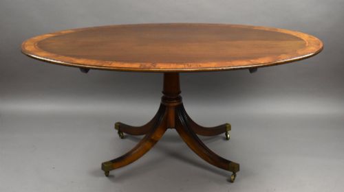 fig 3. An oval Regency style flame mahogany tilt-top dining table featuring a crossbanded satinwood border and a pedestal base with brass castors.
