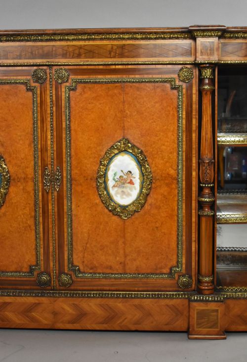 fig 3. A Victorian burr walnut credenza featuring central cupboard doors with oval porcelain plaques and gilt bronze mounts flanked by curved open display shelves.
