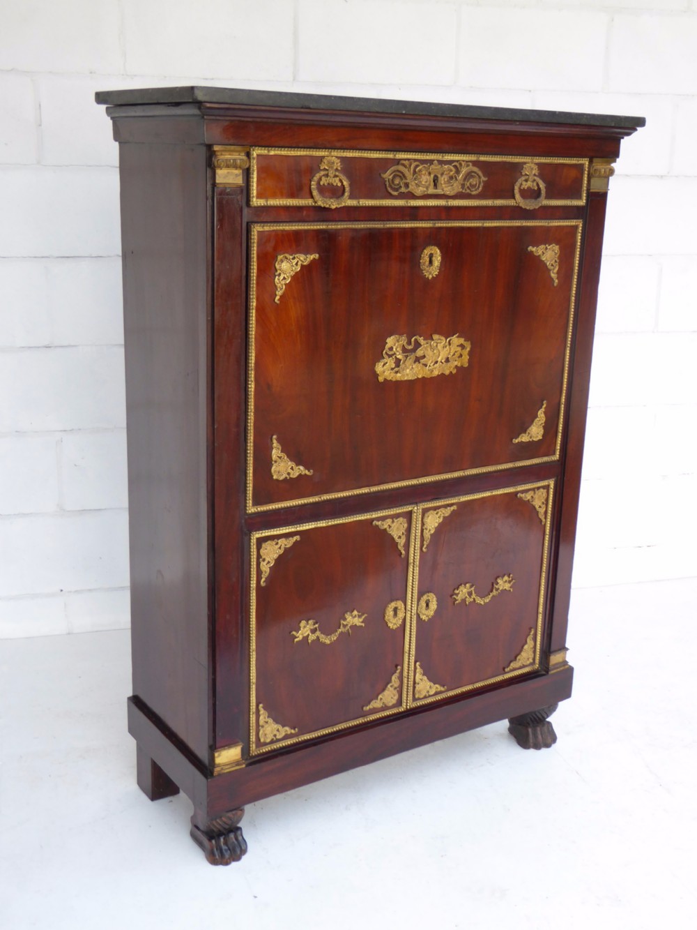 A dark mahogany drop-front escritoire cabinet featuring an open writing flap, fitted interior drawers, brass mounts, a black marble top, and carved paw feet. View II