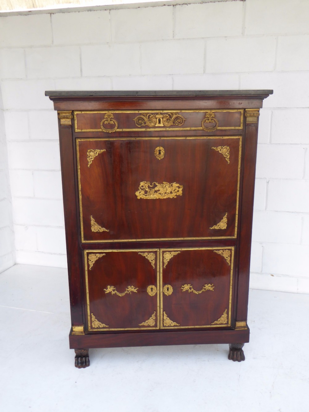 A dark mahogany drop-front escritoire cabinet featuring an open writing flap, fitted interior drawers, brass mounts, a black marble top, and carved paw feet. View III