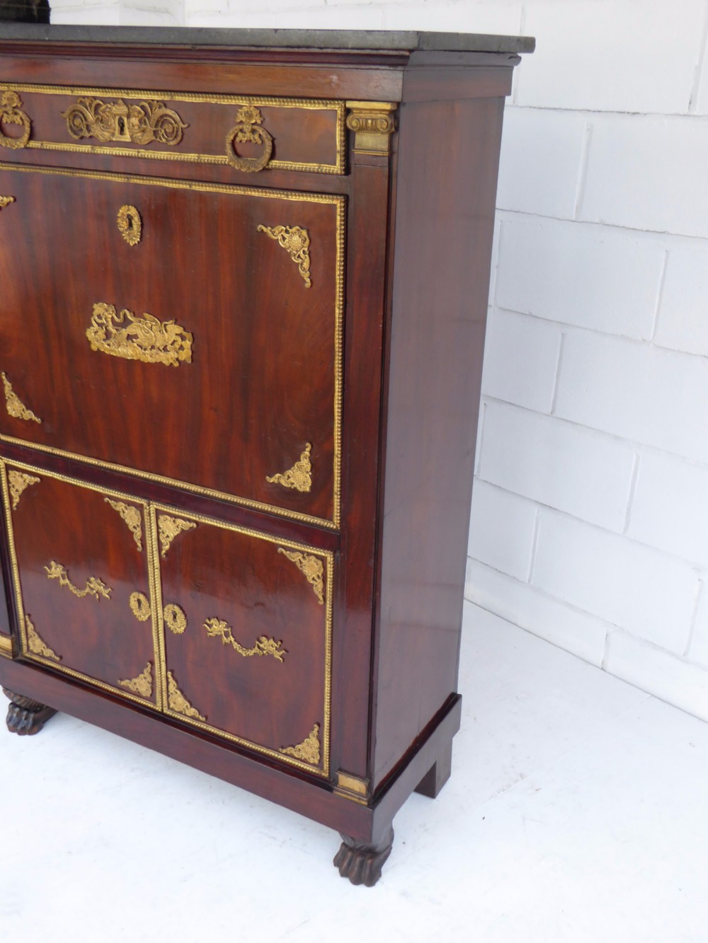 A dark mahogany drop-front escritoire cabinet featuring an open writing flap, fitted interior drawers, brass mounts, a black marble top, and carved paw feet. View IV