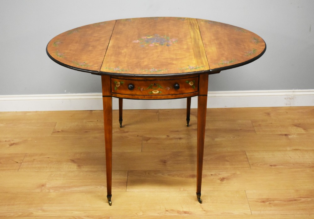 An Edwardian oval drop-leaf Pembroke table crafted from mahogany and satinwood, decorated with painted floral motifs and supported on tapered legs with brass castors.