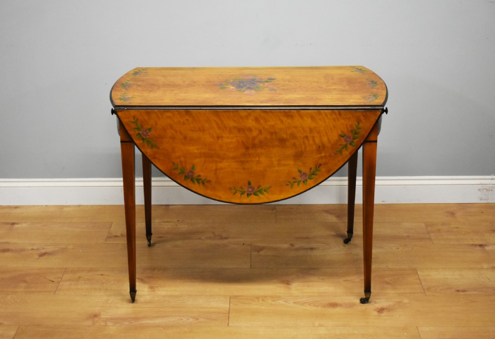 An Edwardian oval drop-leaf Pembroke table crafted from mahogany and satinwood, decorated with painted floral motifs and supported on tapered legs with brass castors. View III