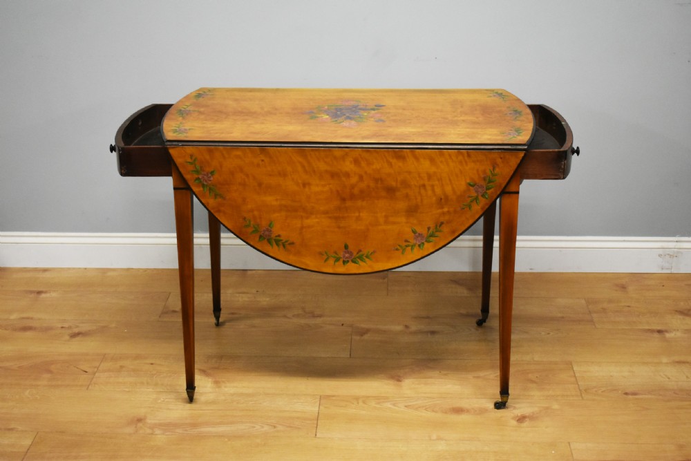 An Edwardian oval drop-leaf Pembroke table crafted from mahogany and satinwood, decorated with painted floral motifs and supported on tapered legs with brass castors. View IV