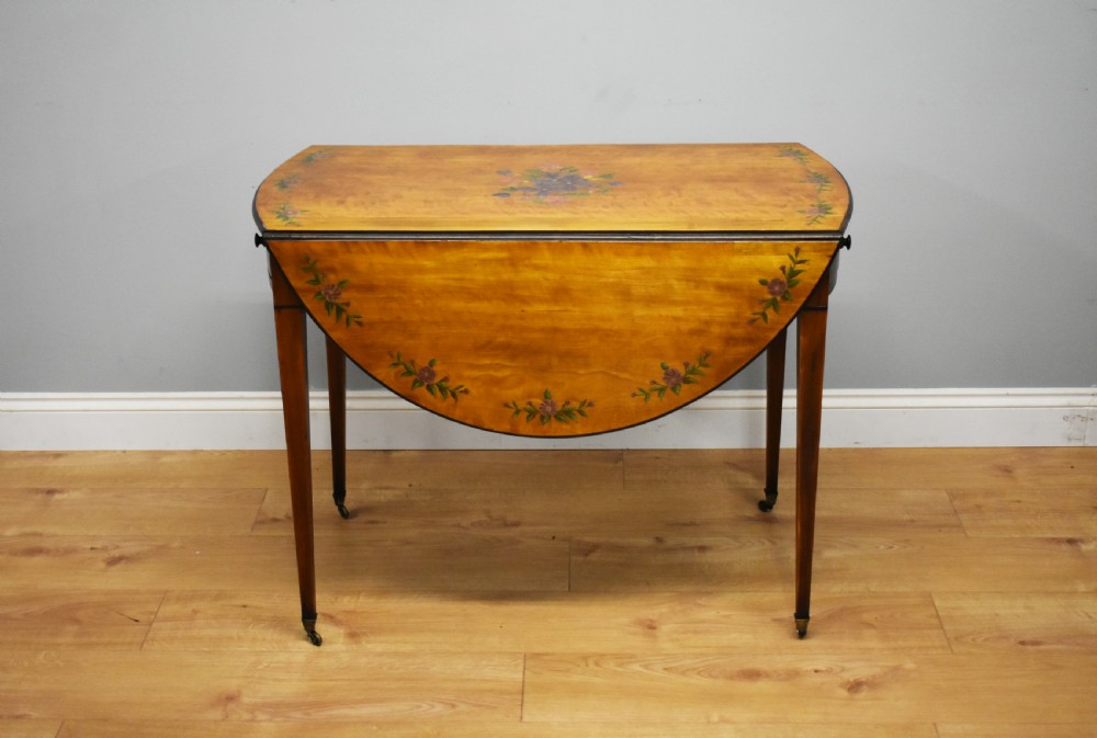 An Edwardian oval drop-leaf Pembroke table crafted from mahogany and satinwood, decorated with painted floral motifs and supported on tapered legs with brass castors. View V