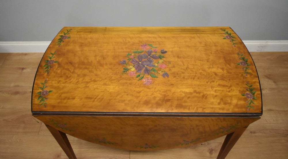 An Edwardian oval drop-leaf Pembroke table crafted from mahogany and satinwood, decorated with painted floral motifs and supported on tapered legs with brass castors. View VI
