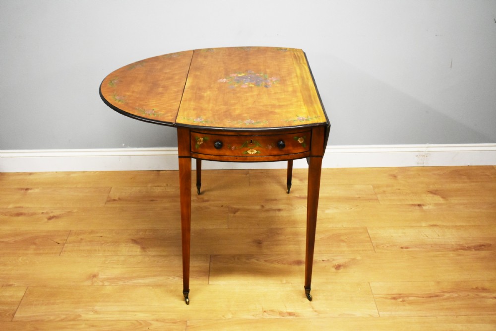 An Edwardian oval drop-leaf Pembroke table crafted from mahogany and satinwood, decorated with painted floral motifs and supported on tapered legs with brass castors. View VII