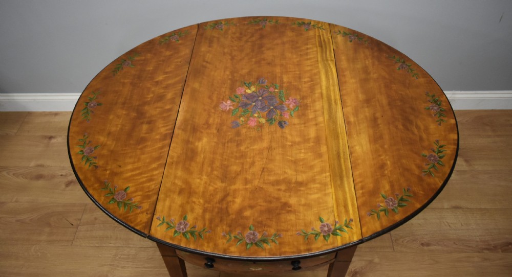 An Edwardian oval drop-leaf Pembroke table crafted from mahogany and satinwood, decorated with painted floral motifs and supported on tapered legs with brass castors. View VIII