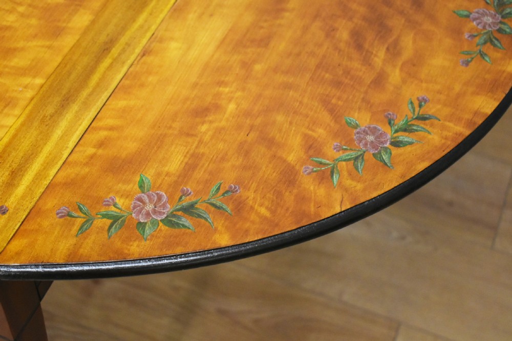 An Edwardian oval drop-leaf Pembroke table crafted from mahogany and satinwood, decorated with painted floral motifs and supported on tapered legs with brass castors. View X