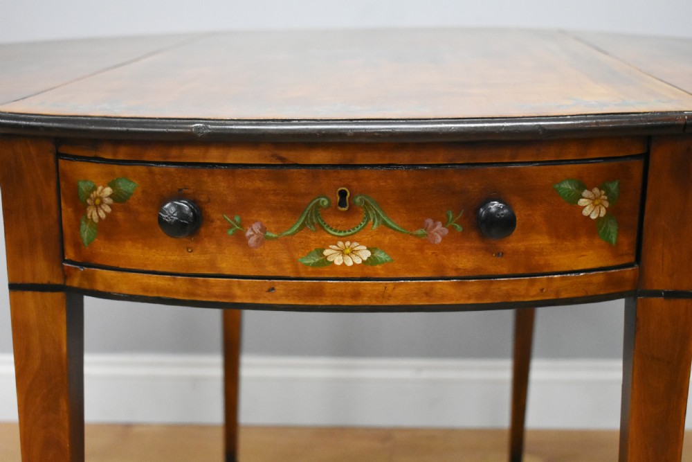 An Edwardian oval drop-leaf Pembroke table crafted from mahogany and satinwood, decorated with painted floral motifs and supported on tapered legs with brass castors. View XI