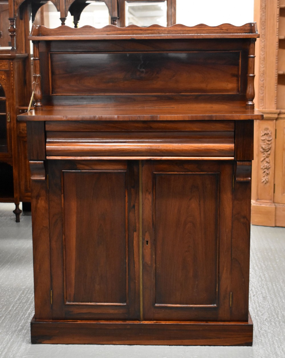 A Victorian rosewood chiffonier cabinet featuring twin panelled doors, a long frieze drawer, an upper gallery shelf with turned supports, and a solid plinth base.