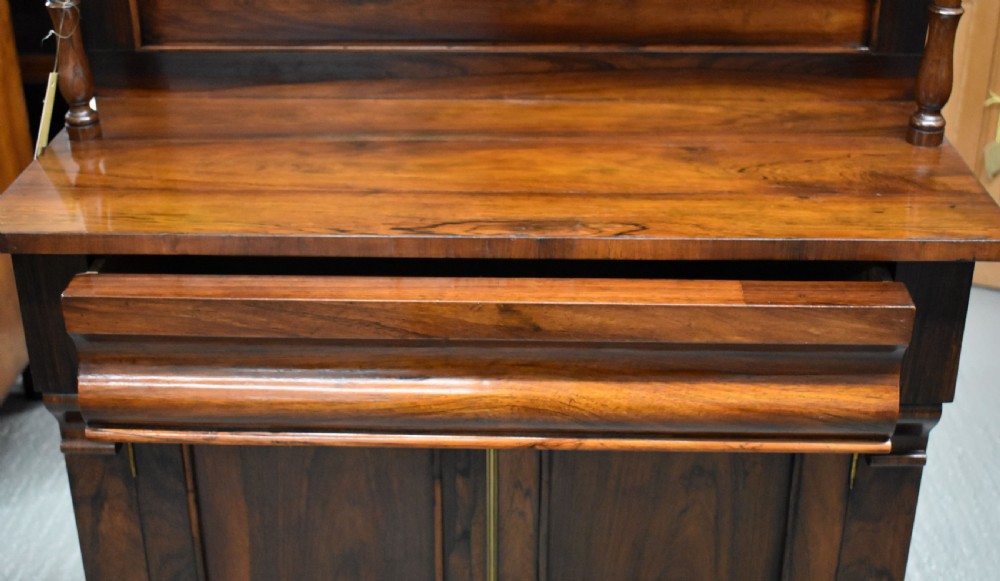 A Victorian rosewood chiffonier cabinet featuring twin panelled doors, a long frieze drawer, an upper gallery shelf with turned supports, and a solid plinth base. View VI