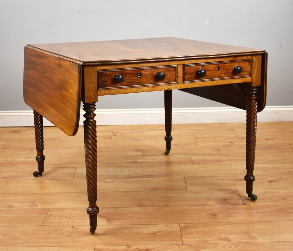 A Regency period mahogany drop-leaf table featuring two drawers with dark turned knobs and four spiral-carved legs on brass castors.