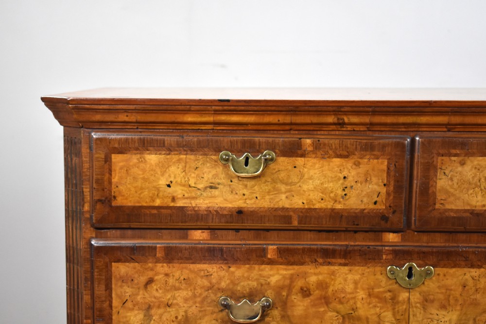 A George III burr walnut chest of drawers featuring two short drawers over three long drawers with crossbanded borders, brass escutcheons, and bun feet. View VII