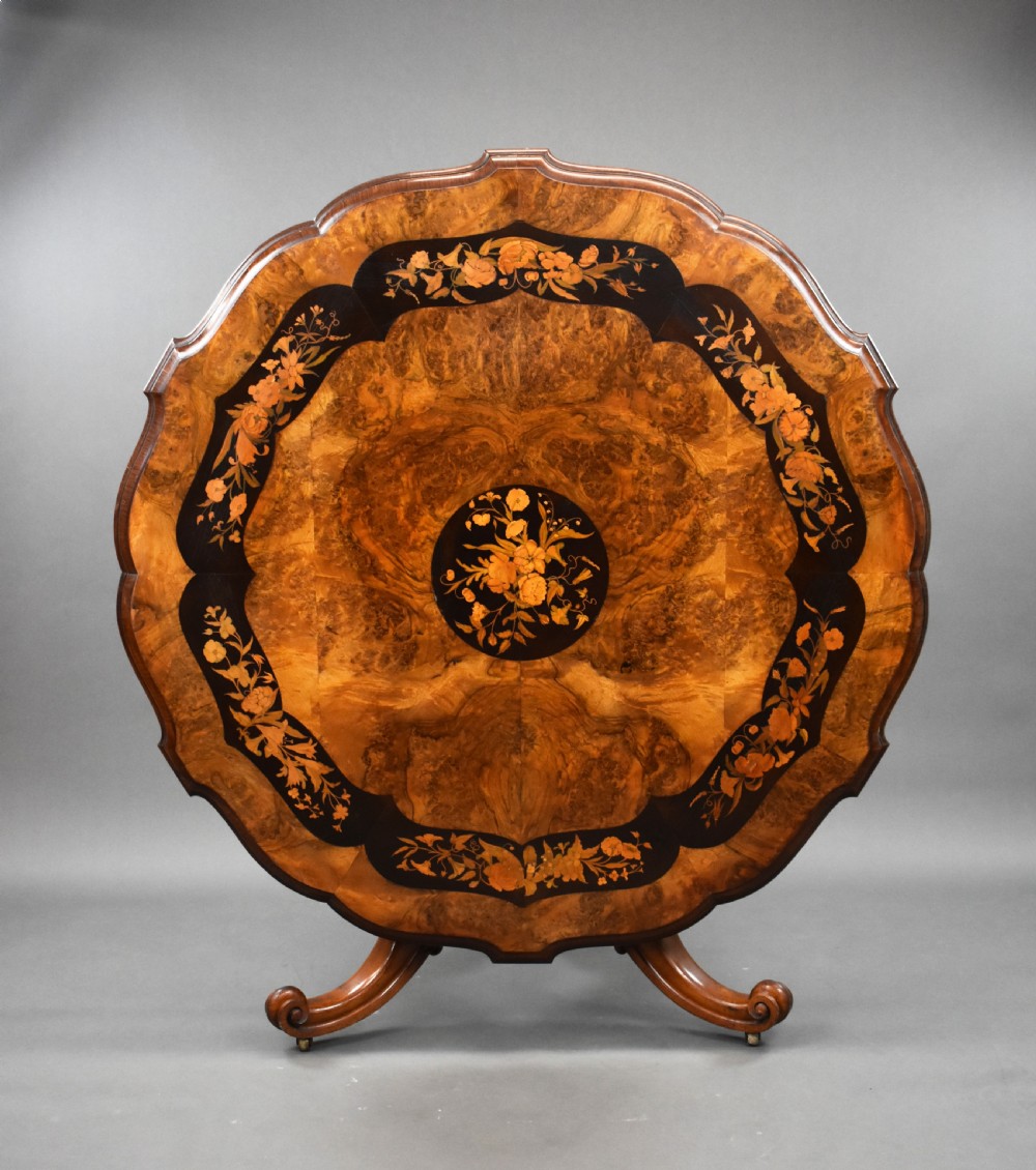 A Victorian burr walnut tilt-top breakfast table featuring a scalloped edge, intricate floral marquetry inlay border, and a carved four-star pedestal base.