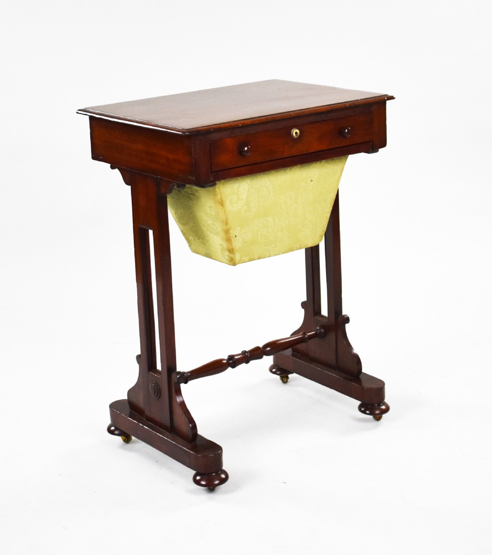 A Victorian mahogany work table featuring a single drawer with wooden knobs, a suspended yellow patterned fabric basket below, and a turned stretcher base set on castors.