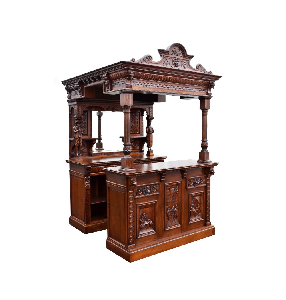 A large, ornate carved oak front and back bar unit featuring detailed figural panels, turned columns, and an overhead canopy with a swan neck pediment.