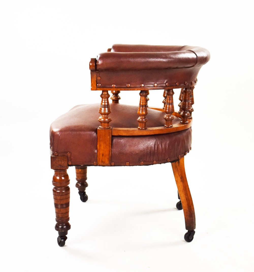 A Victorian oak tub armchair featuring brown leather upholstery, turned wooden spindles, stud detailing, and legs set on castors. View V