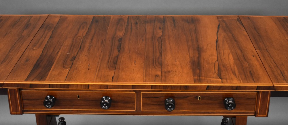 An early 19th-century Regency rosewood sofa table with drop leaves, boxwood stringing, two drawers, turned legs, and brass castors. View X