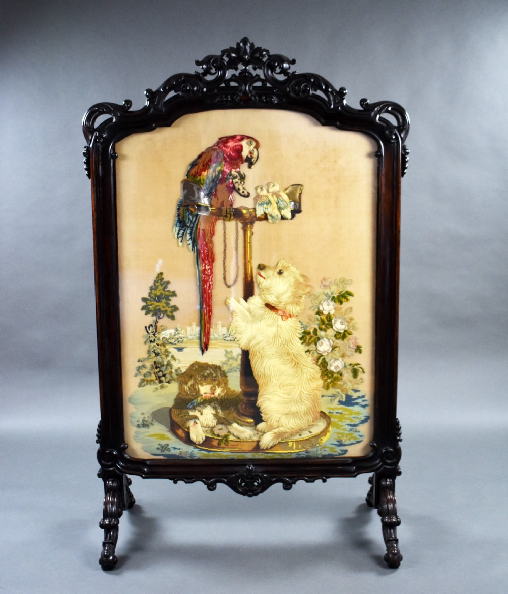 A dark carved wood Victorian fire screen framed around a central needlepoint panel depicting a parrot perched above two dogs.