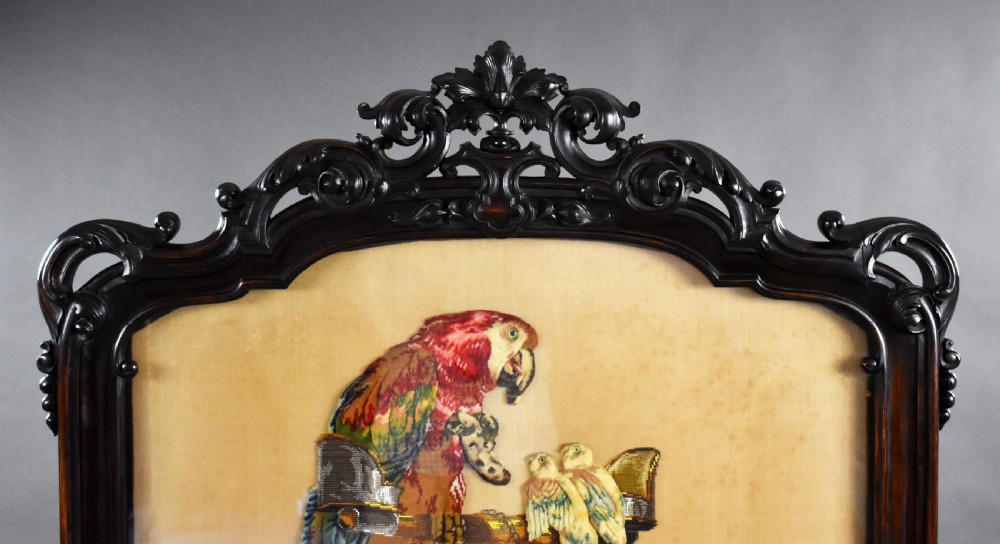 A dark carved wood Victorian fire screen framed around a central needlepoint panel depicting a parrot perched above two dogs. View II