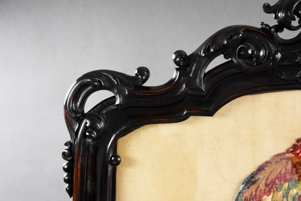 A dark carved wood Victorian fire screen framed around a central needlepoint panel depicting a parrot perched above two dogs. View III