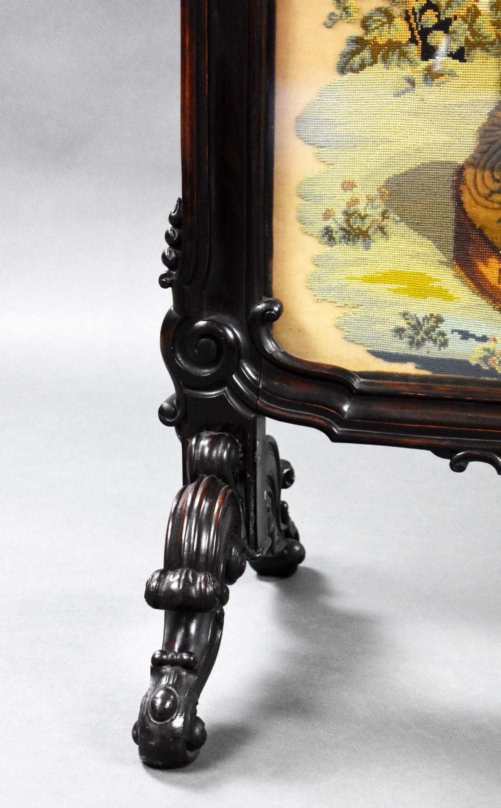 A dark carved wood Victorian fire screen framed around a central needlepoint panel depicting a parrot perched above two dogs. View VII