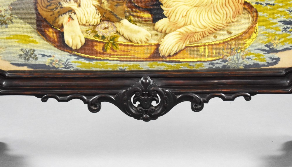 A dark carved wood Victorian fire screen framed around a central needlepoint panel depicting a parrot perched above two dogs. View VIII