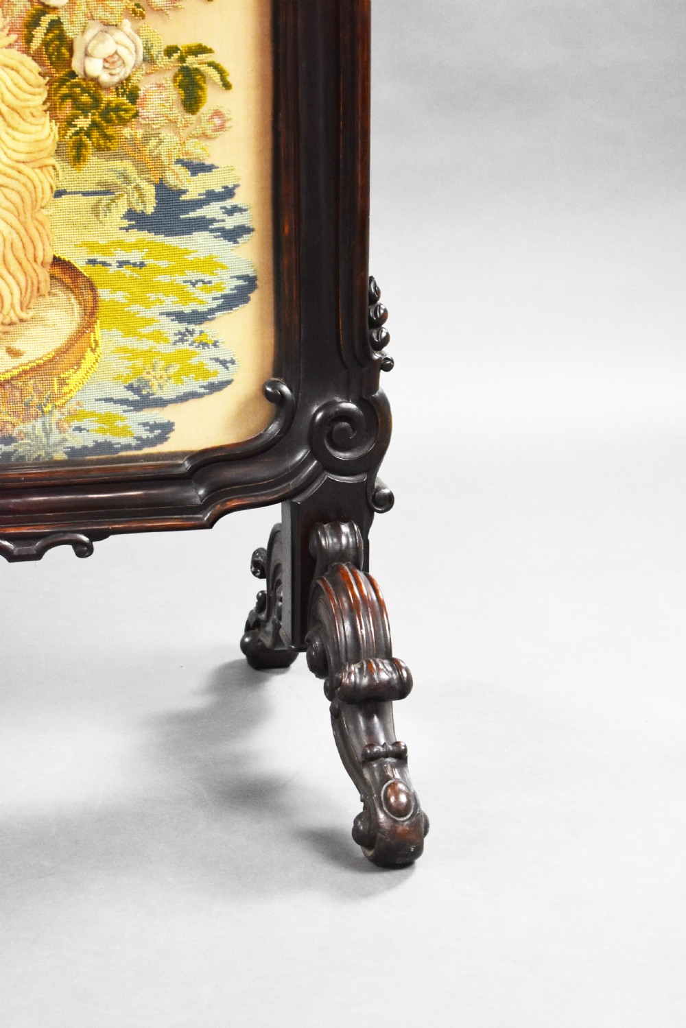 A dark carved wood Victorian fire screen framed around a central needlepoint panel depicting a parrot perched above two dogs. View IX