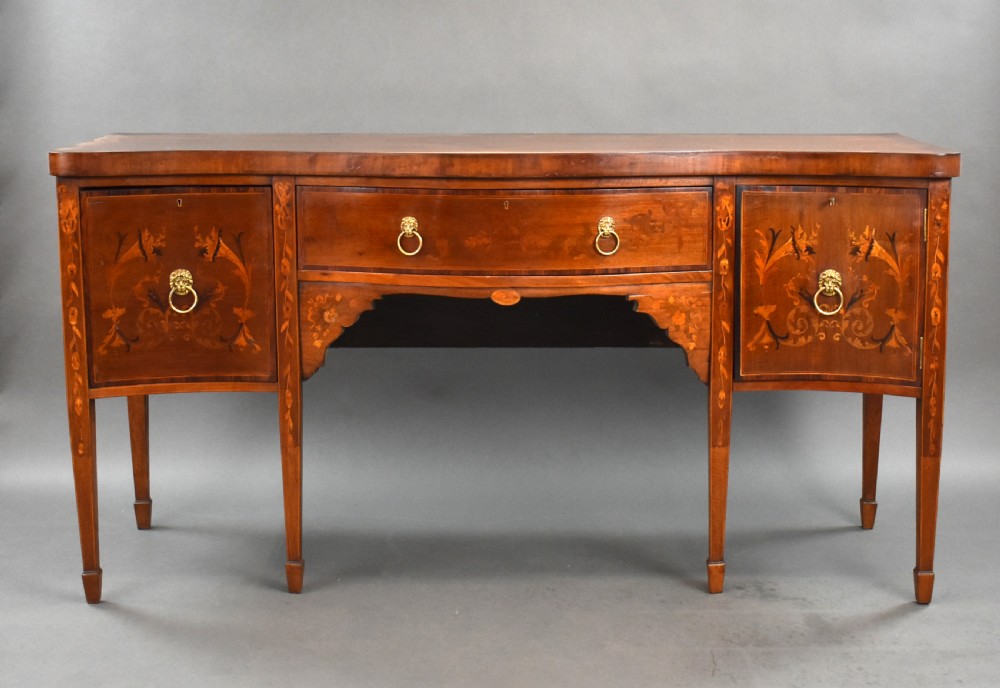 An Edwardian mahogany serpentine sideboard featuring satinwood marquetry inlay, brass lion mask ring handles, and tapered square legs.