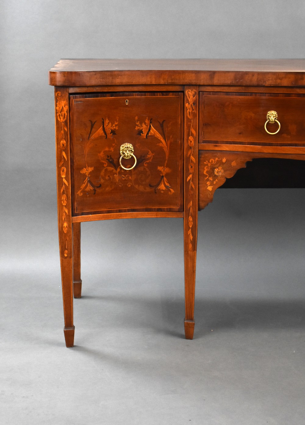 An Edwardian mahogany serpentine sideboard featuring satinwood marquetry inlay, brass lion mask ring handles, and tapered square legs. View II