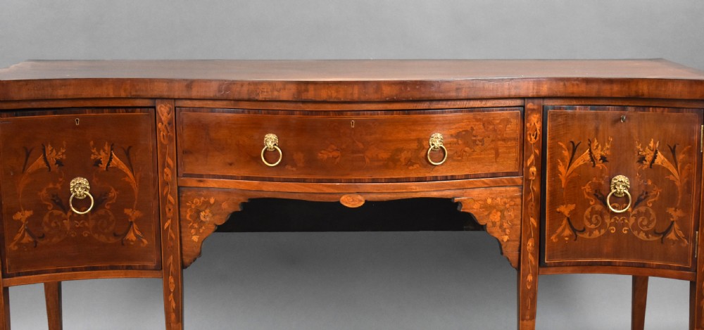An Edwardian mahogany serpentine sideboard featuring satinwood marquetry inlay, brass lion mask ring handles, and tapered square legs. View III