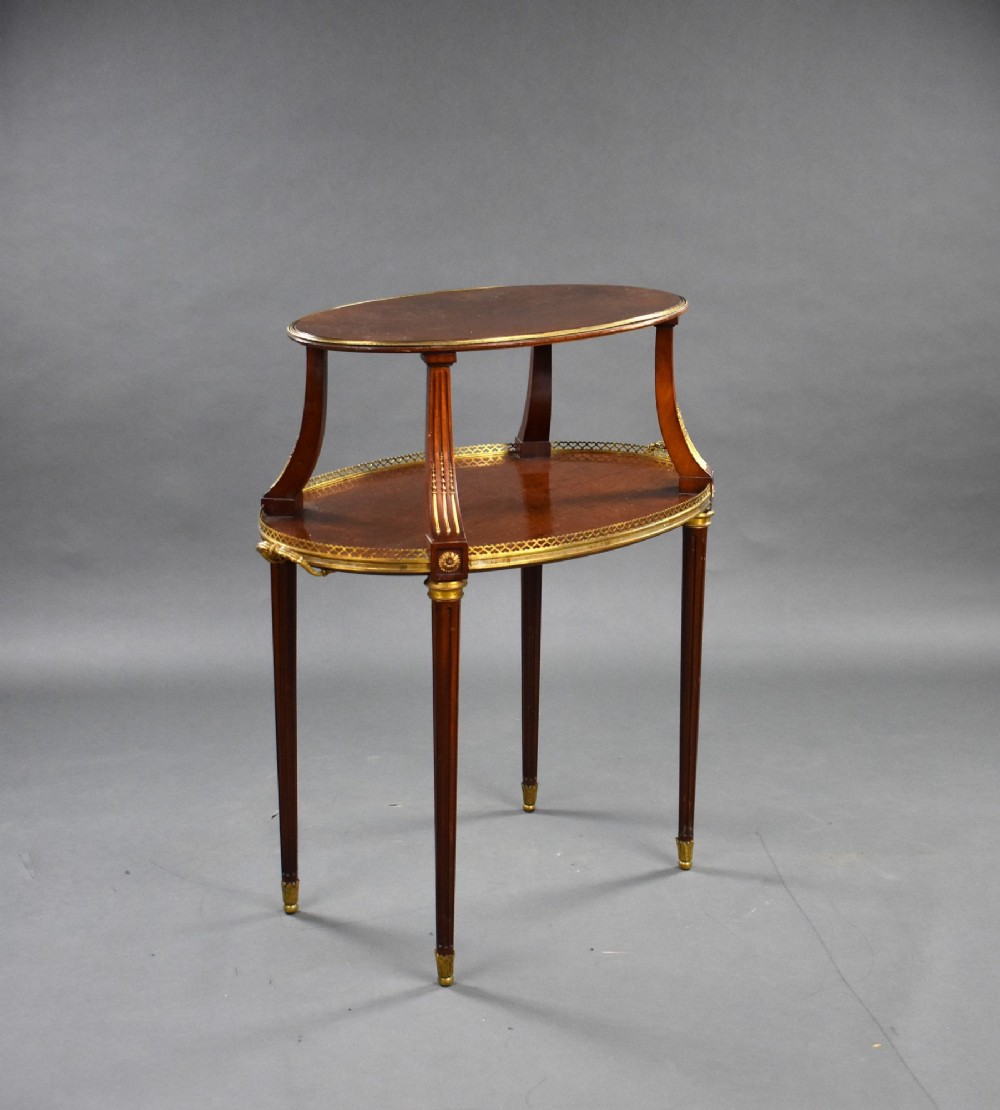 A two-tiered oval French mahogany etagere table featuring gilt-bronze mounts, a pierced brass gallery shelf, and slim fluted legs with brass feet.