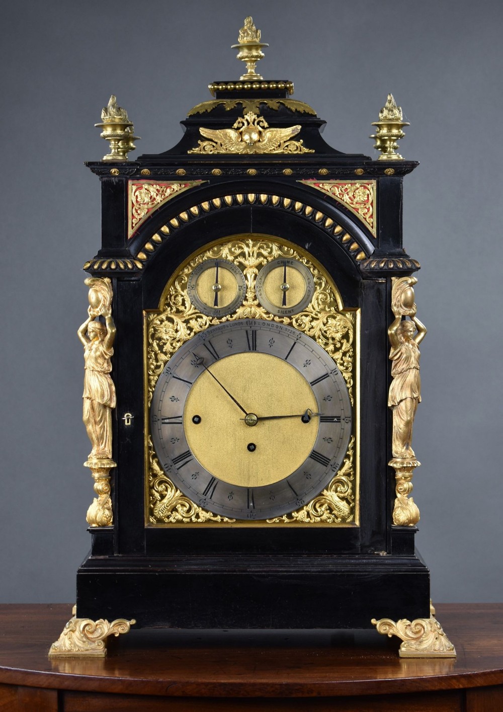 A Victorian ebonised bracket clock featuring an arched black wooden case, gilded brass caryatids, a gold spandrel dial with Roman numerals, and top flame finials.