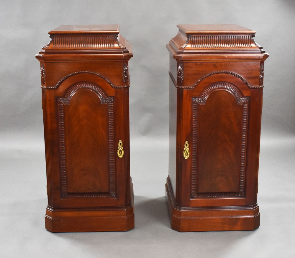 A matching pair of tall 19th-century mahogany pedestal cabinets featuring arched carved doors, fluted top mouldings, and brass drop handles.