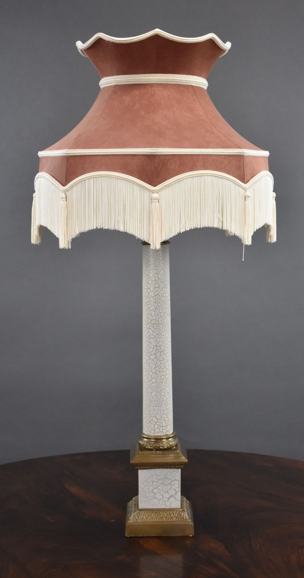 An Empire style table lamp featuring a crackle-glaze column on a stepped brass base, topped with a terracotta fabric shade trimmed with cream braid and fringe.