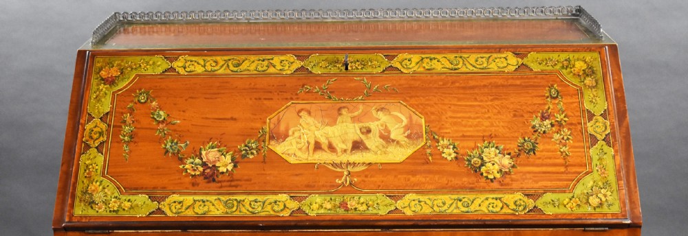 An Edwardian hand-painted satinwood bureau featuring a slant front with a classical central medallion and floral swags above four graduated drawers. View X