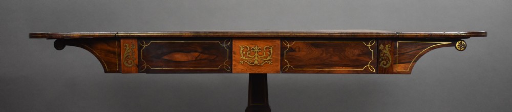 A Regency period dark rosewood sofa table with intricate brass inlay, two drop leaves, a central pedestal support, and four splayed legs with brass claw castors. View VIII