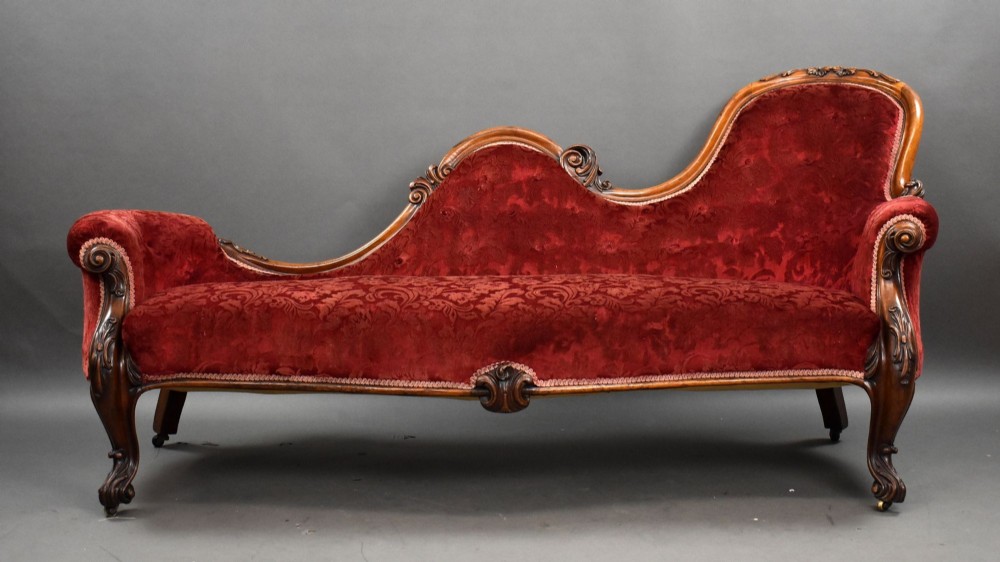 A Victorian mahogany chaise longue upholstered in patterned deep red velvet featuring an asymmetric scrolled backrest and cabriole legs.
