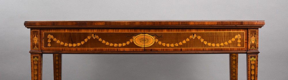 A 19th-century rectangular wooden writing desk featuring detailed marquetry swags on the apron, a leather writing surface, and four square tapered legs on brass castors. View VI