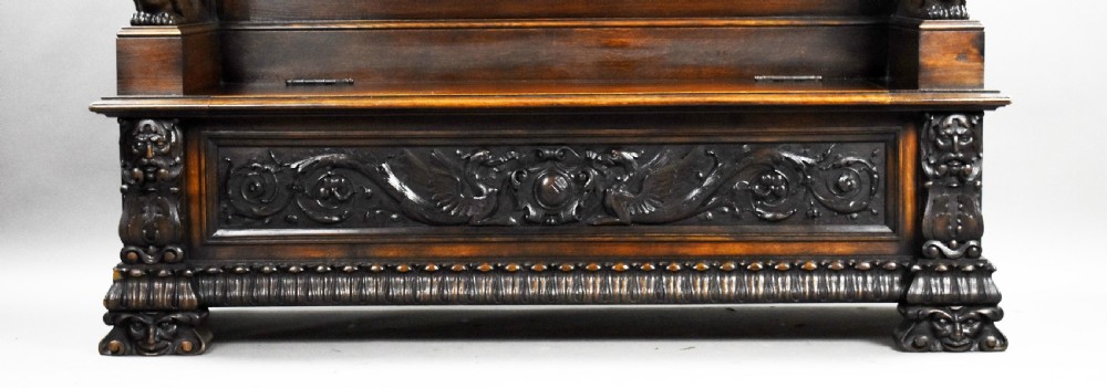 A dark carved oak hall bench featuring a high ornate backrest with a heraldic crest, carved mythic figure armrests, and a hinged chest seat. View VI