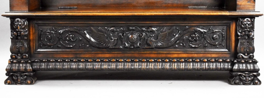 A dark carved oak hall bench featuring a high ornate backrest with a heraldic crest, carved mythic figure armrests, and a hinged chest seat. View XIV