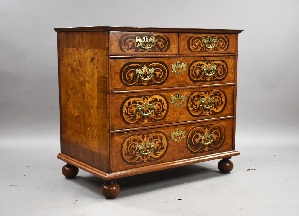 A William and Mary burr walnut chest featuring intricate dark marquetry inlay panels, brass swing handles, and turned bun feet.